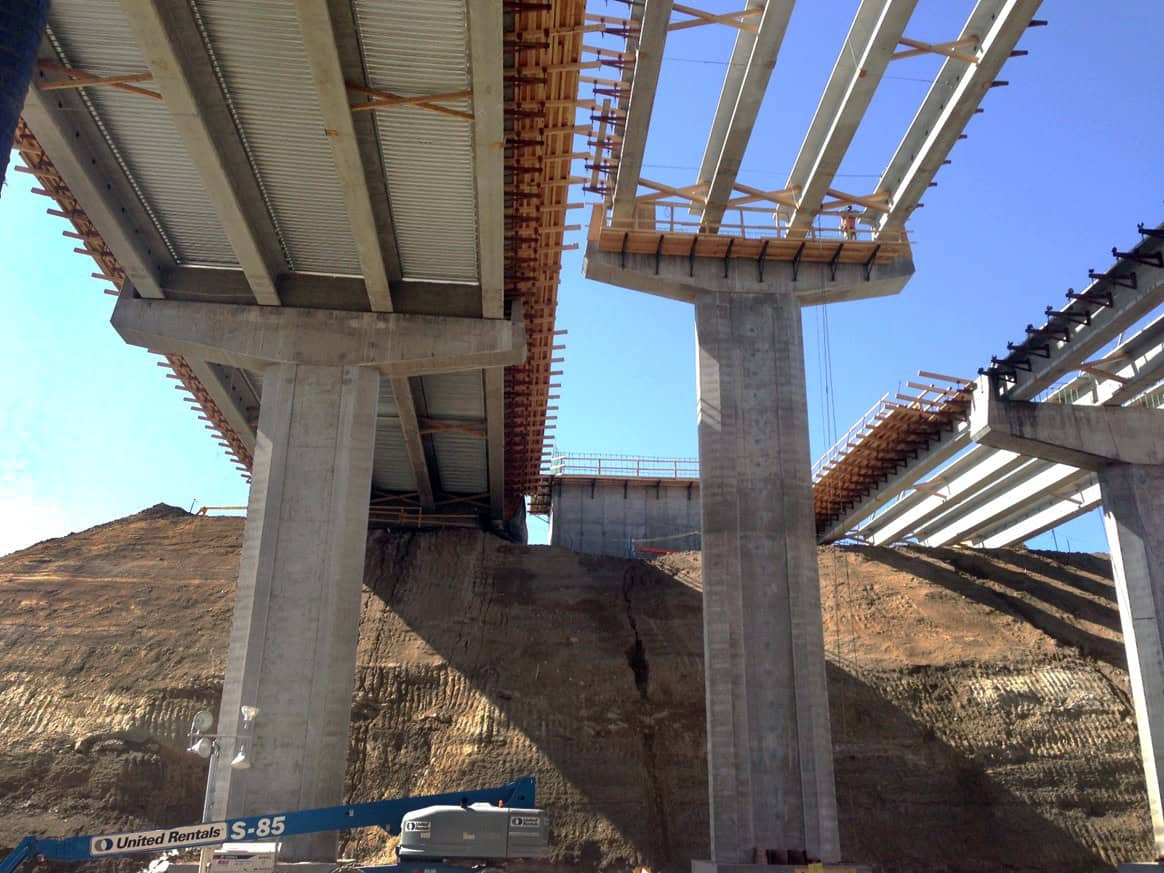 Concrete bridge pillars supporting partially built spans while construction equipment works at their base on a terraced dirt hillside under a clear blue sky.

Text in image: "United Rentals S-85"