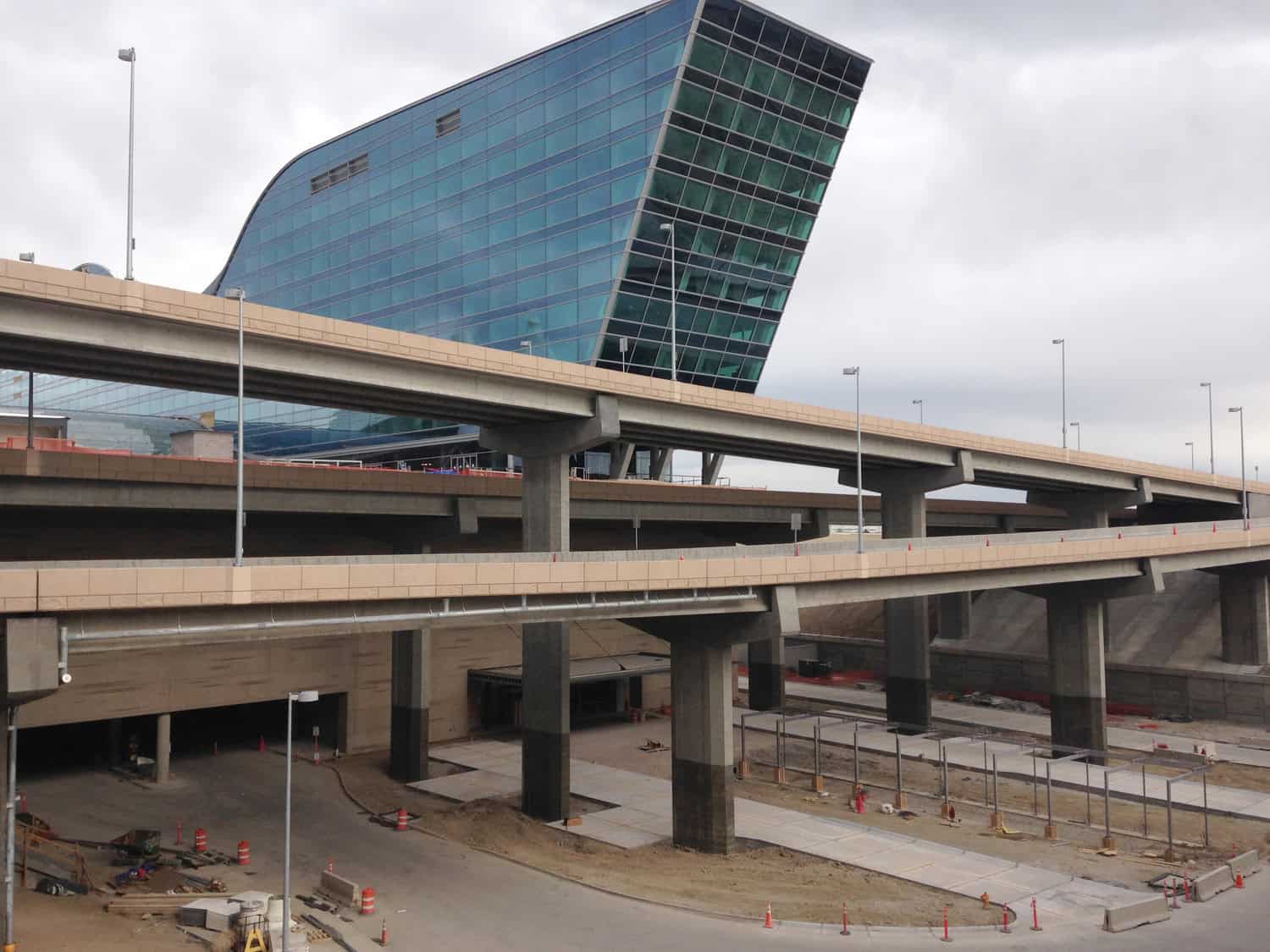 Glass-paneled slanted office tower leans behind layered concrete highway ramps; ramps cross and curve above a construction site with traffic cones, barriers, and unfinished pavement under an overcast sky.