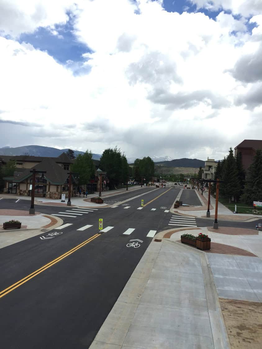 A wide, empty downtown street stretches toward distant snow‑capped mountains; painted bike lanes and crosswalks guide traffic while shops, trees, lamp posts, and planters flank sidewalks beneath a cloudy sky.