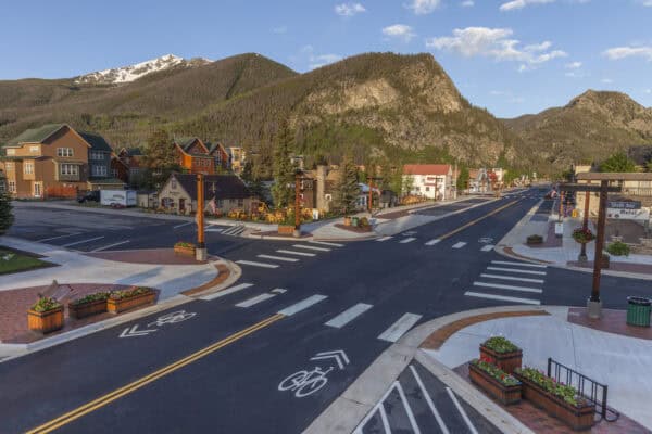 Main street stretches empty through a small mountain town, flanked by shops, planters, crosswalks and bike lanes, with forested cliffs and a snow‑capped peak rising behind.

Text in image: "Sixth Ave Motel"; "ANTIQUES"