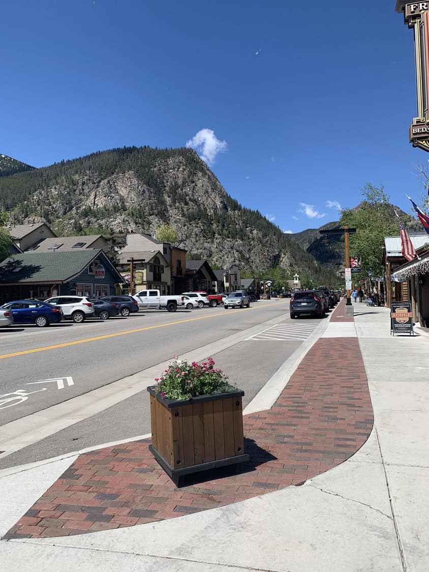 A wooden planter of pink flowers sits on a brick strip beside a sidewalk; a mountain town street with parked cars, storefronts, and a rocky forested peak under a clear blue sky.

Text visible: "LODGE" (other signs present but not legible)