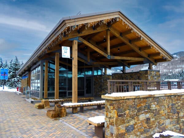 Sheltered wooden bus stop stands empty beneath a large peaked timber roof; snow-topped benches and stone walls sit on a paved plaza with snowy trees and mountains beyond. Text: "BUS STOP" (bus icon), "P", "eco".