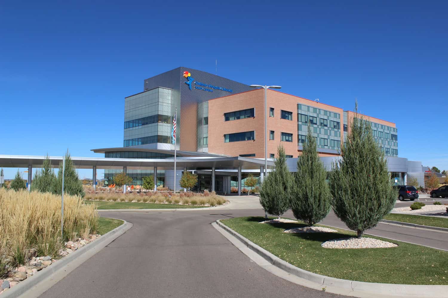A multi-story hospital building (Children's Hospital Colorado) fronts a covered drop-off and circular driveway; landscaped islands with conifer trees and ornamental grasses sit beneath a clear blue sky.