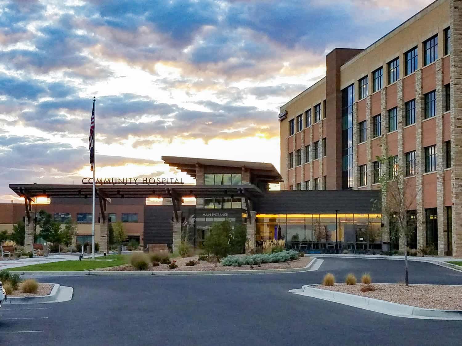 Community hospital building stands beneath a colorful sunset; covered entrance canopy shelters the main entry, with a flagpole, landscaped forecourt and empty parking lot in front.

Text visible: "COMMUNITY HOSPITAL"; "MAIN ENTRANCE"