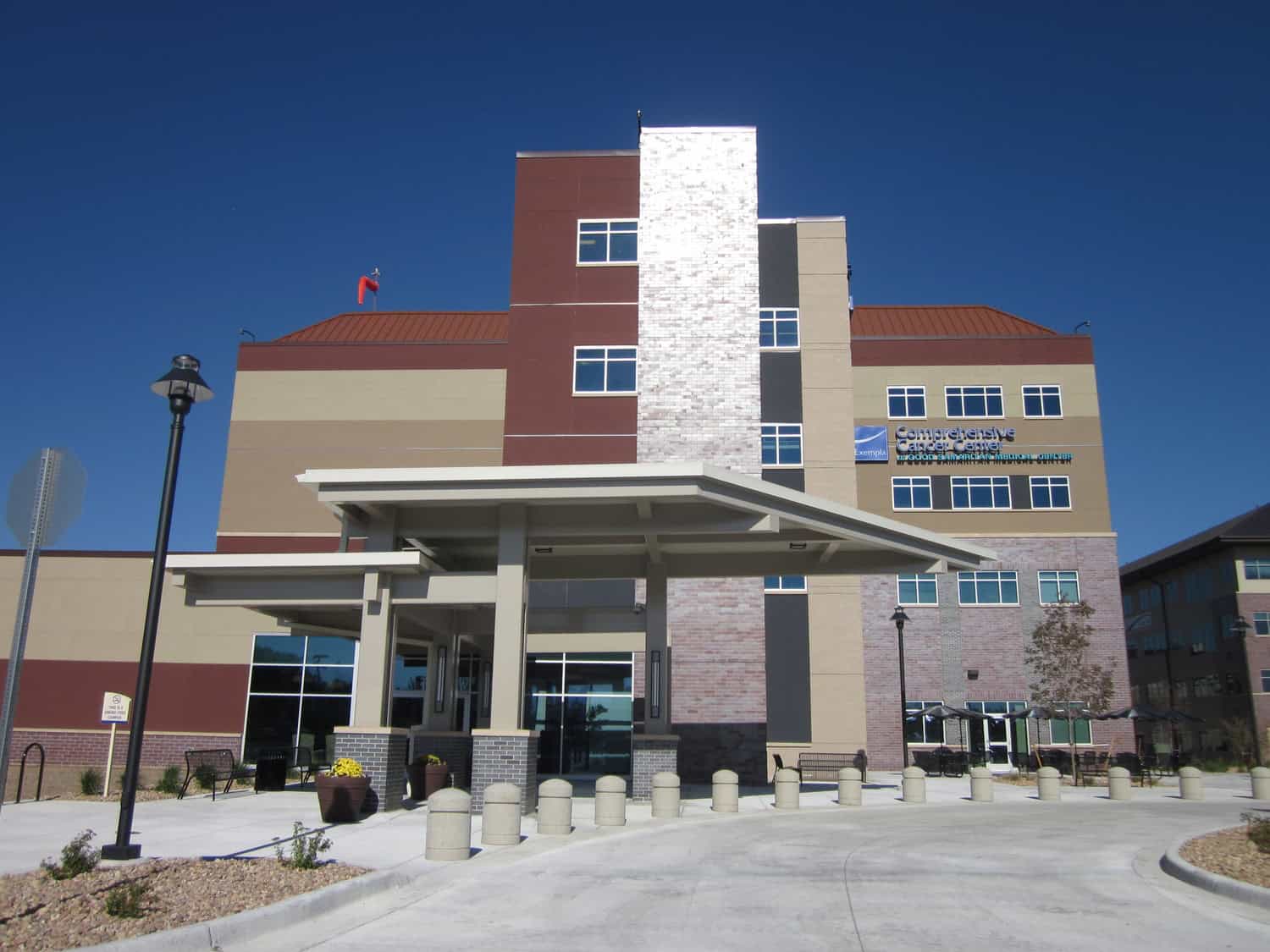 Main hospital entrance canopy shelters a drop-off lane while bollards, planters, and benches sit before a multi-story brick-and-stucco building under a clear blue sky.

Text in image:
"Exempla
Comprehensive Cancer Center
Good Samaritan Medical Center"