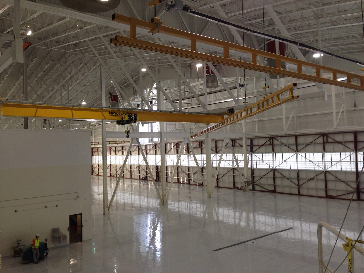 Yellow overhead crane spans the ceiling, hoist suspended above a reflective polished floor inside a vast white aircraft hangar with exposed steel trusses; a worker operates equipment near a doorway.
