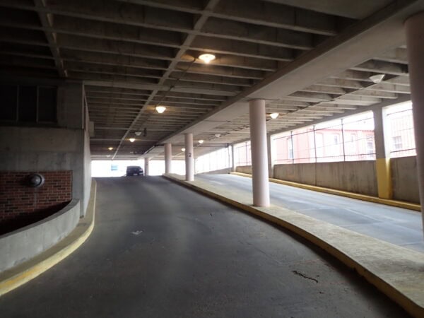 Concrete ramp rises toward a parked car, flanked by pink support columns and yellow curbs inside a dim multi-level parking garage with a grid-pattern concrete ceiling and wall windows.

Text: "2"