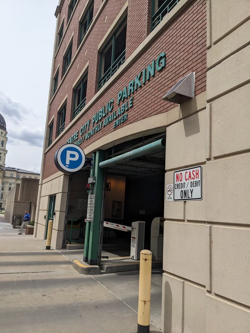 Parking garage entrance (object) with a lowered barrier (action) at a city sidewalk, teal "P" sign and multi-story brick building (context).

Text in image:
- CENTRE CITY PUBLIC PARKING
- HOURLY & MONTHLY AVAILABLE
- ENTER
- (blue circle) P
- NO CASH
- CREDIT / DEBIT ONLY
- NO SMOKING (symbol)