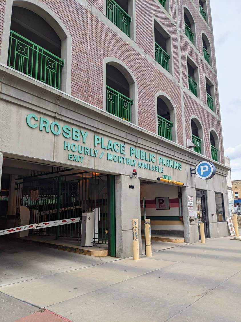 Crosby Place public parking garage facade shows open entry and exit lanes with a raised arm, a round "P" parking sign, and green balcony railings on a multi-story brick building.

Transcribed text:
- CROSBY PLACE PUBLIC PARKING
- HOURLY / MONTHLY AVAILABLE
- EXIT
- ENTER (on yellow clearance bar)
- 106
- (round sign) P