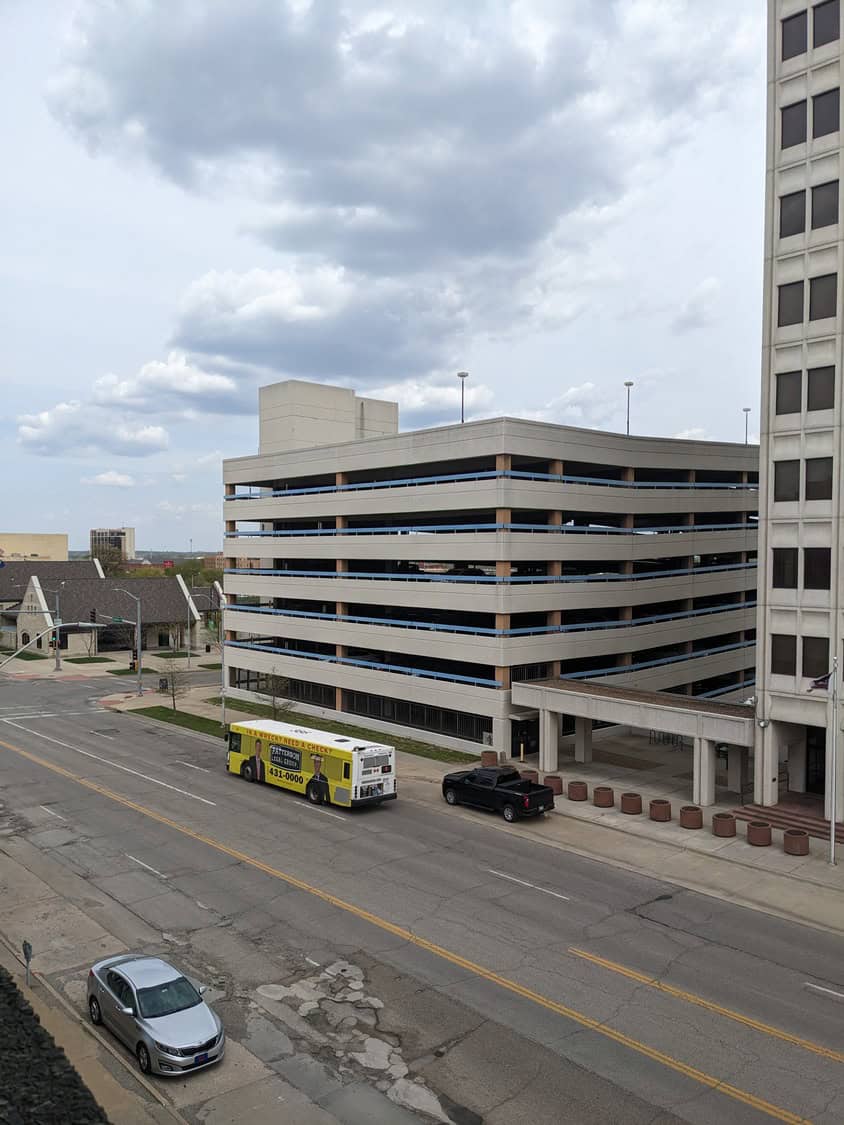 Multi-level parking garage dominates center, flanked by an office tower; a yellow advertising bus drives past while a pickup waits at the entrance, city street and cloudy sky around. Transcribed text: 431-0000; 1000.