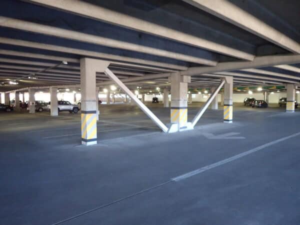 Concrete support columns brace the ceiling with diagonal steel braces, bases wrapped in yellow-striped pads; few cars parked beneath low concrete beams in a dim multi-level parking garage.