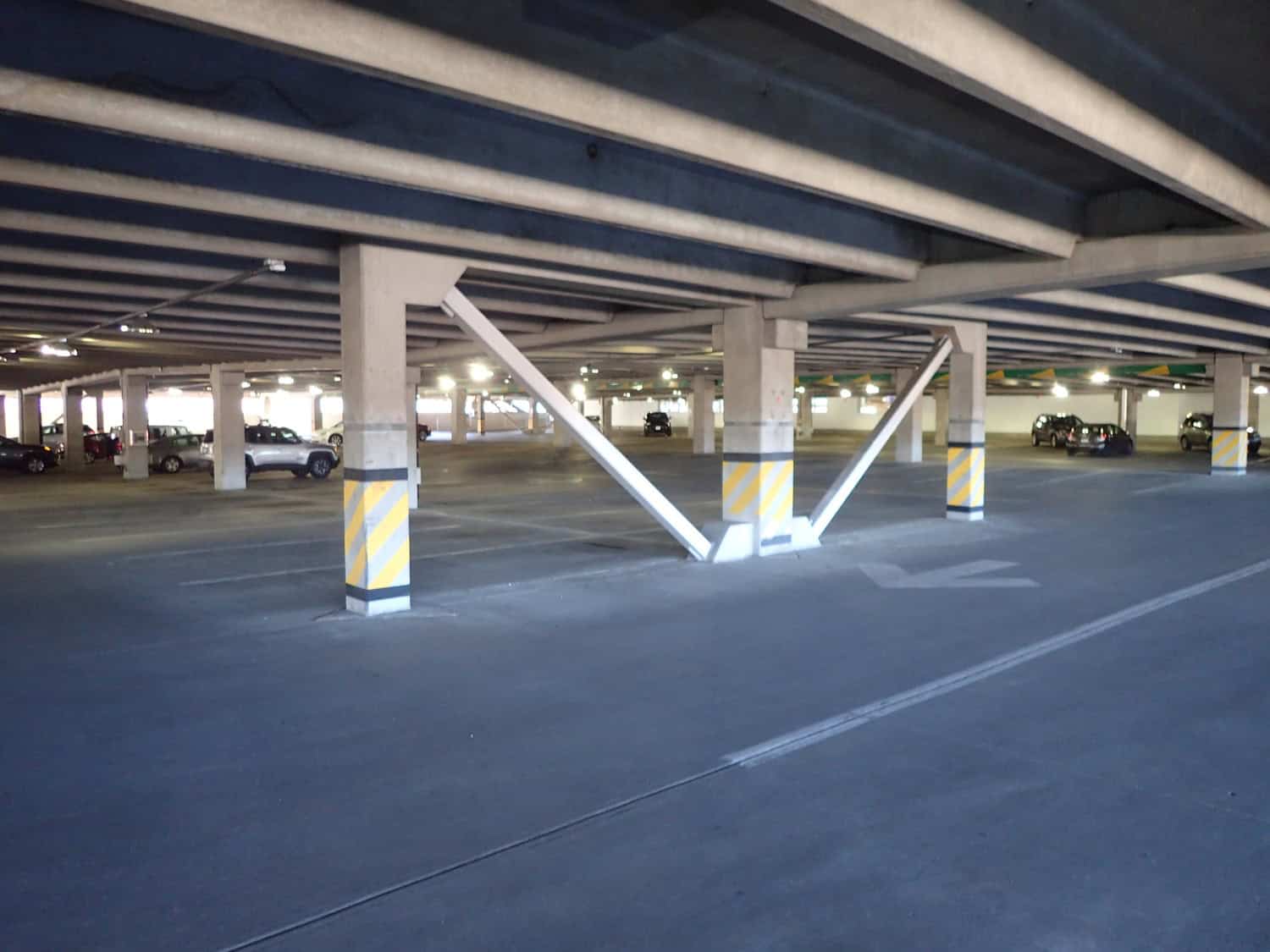 Concrete support columns brace the ceiling with diagonal steel braces, bases wrapped in yellow-striped pads; few cars parked beneath low concrete beams in a dim multi-level parking garage.