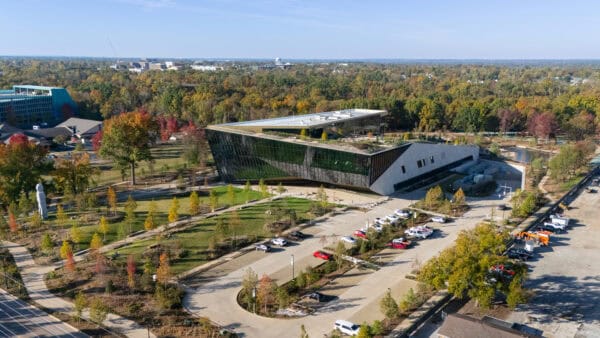An angular glass-and-metal building with a planted green roof sits amid landscaped lawns and tree-lined paths, bordered by parked cars and autumn-colored woods stretching to the horizon under clear sky.