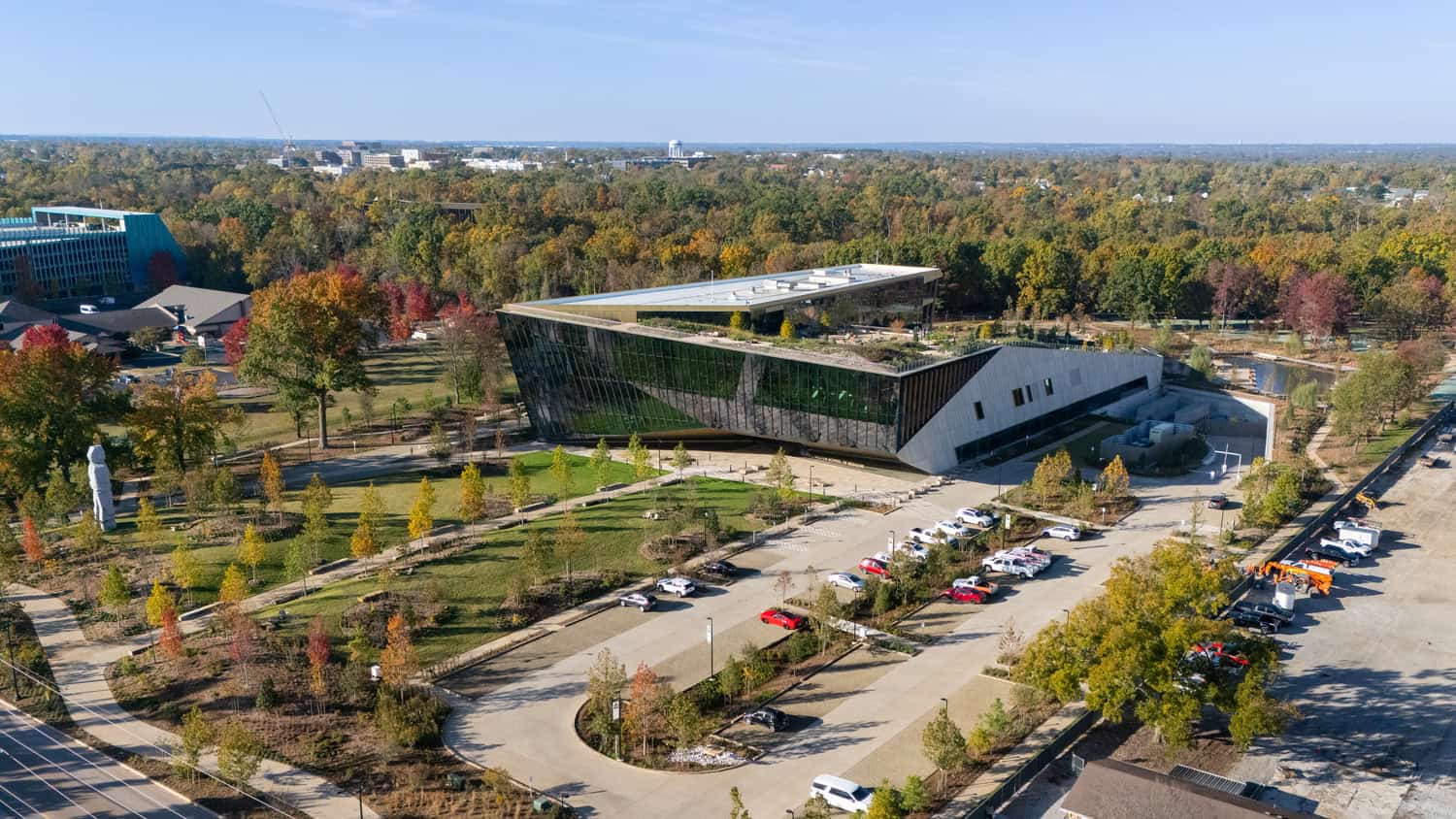 An angular glass-and-metal building with a planted green roof sits amid landscaped lawns and tree-lined paths, bordered by parked cars and autumn-colored woods stretching to the horizon under clear sky.