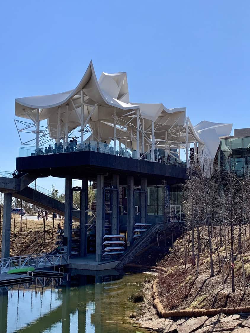 White undulating-roof pavilion perched on concrete stilts, hosting diners on a glass-railed terrace above a narrow pond with kayaks stored beneath and young trees lining the bank.