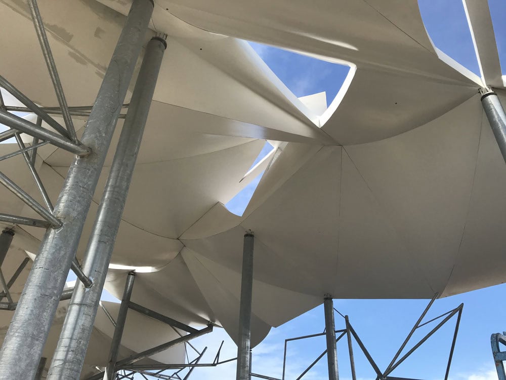 White tensile fabric canopy stretching and folding into pointed, star-shaped openings, supported by galvanized steel poles and scaffolding, viewed from below against a clear blue sky.