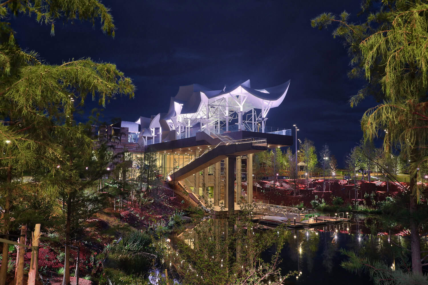 White, sail-like pavilion sits illuminated atop concrete columns, casting reflections on a calm pond, connected by a staircase and docks within a landscaped, tree-lined park at night.