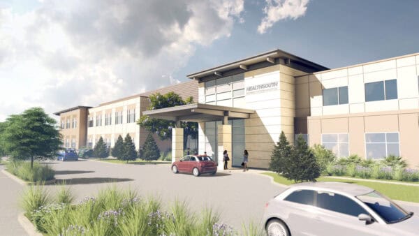 Sign on façade: HEALTHSOUTH REHABILITATION HOSPITAL. Two-story hospital building stands with a covered drop-off (porte-cochère) where people walk and cars pull up; landscaped grounds and cloudy sky.