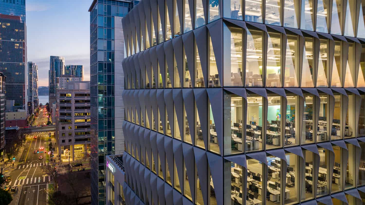 Office tower — illuminated, empty open‑plan workspaces visible through a repeating angular glass facade; downtown high-rise street extends toward a waterfront at dusk.