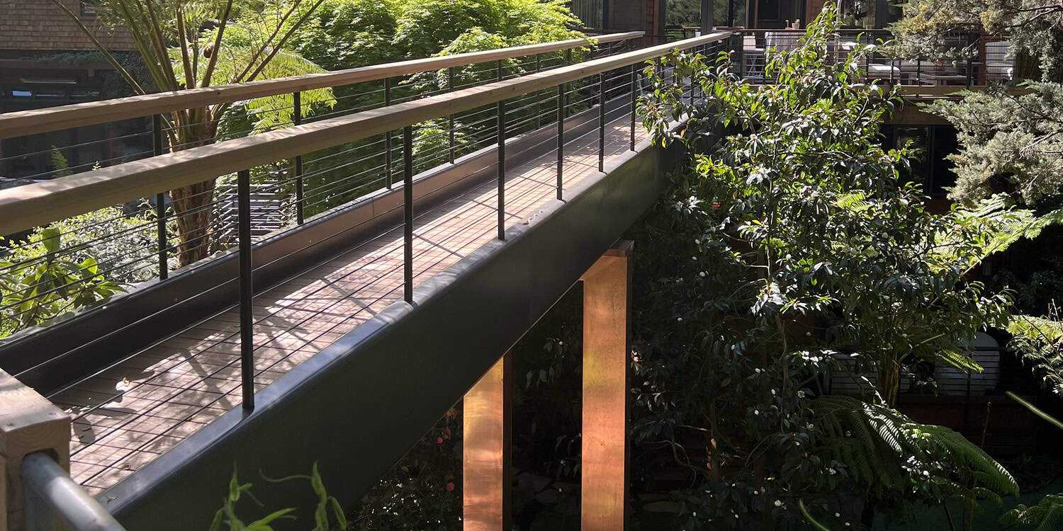 A raised wooden walkway with metal railings crosses over a lush indoor courtyard; sunlight casts shadows on the deck while copper-clad support columns rise amid dense tropical plants.