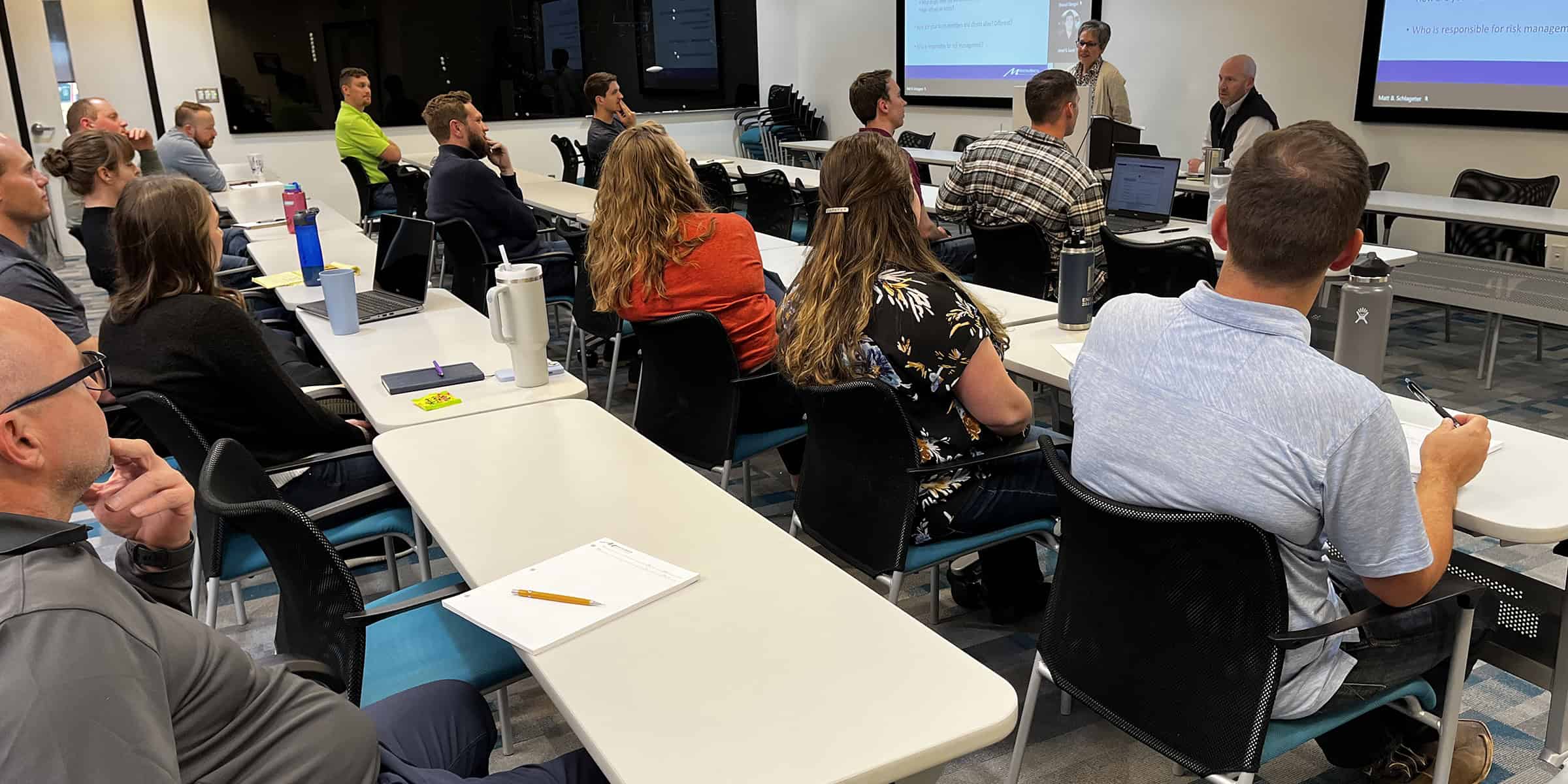 Classroom of adults (attendees) listening to two presenters at the front; people seated at tables with laptops, notebooks, and water bottles; projection screens and a lectern in a modern meeting room. Projection-screen text is too small and illegible to transcribe.