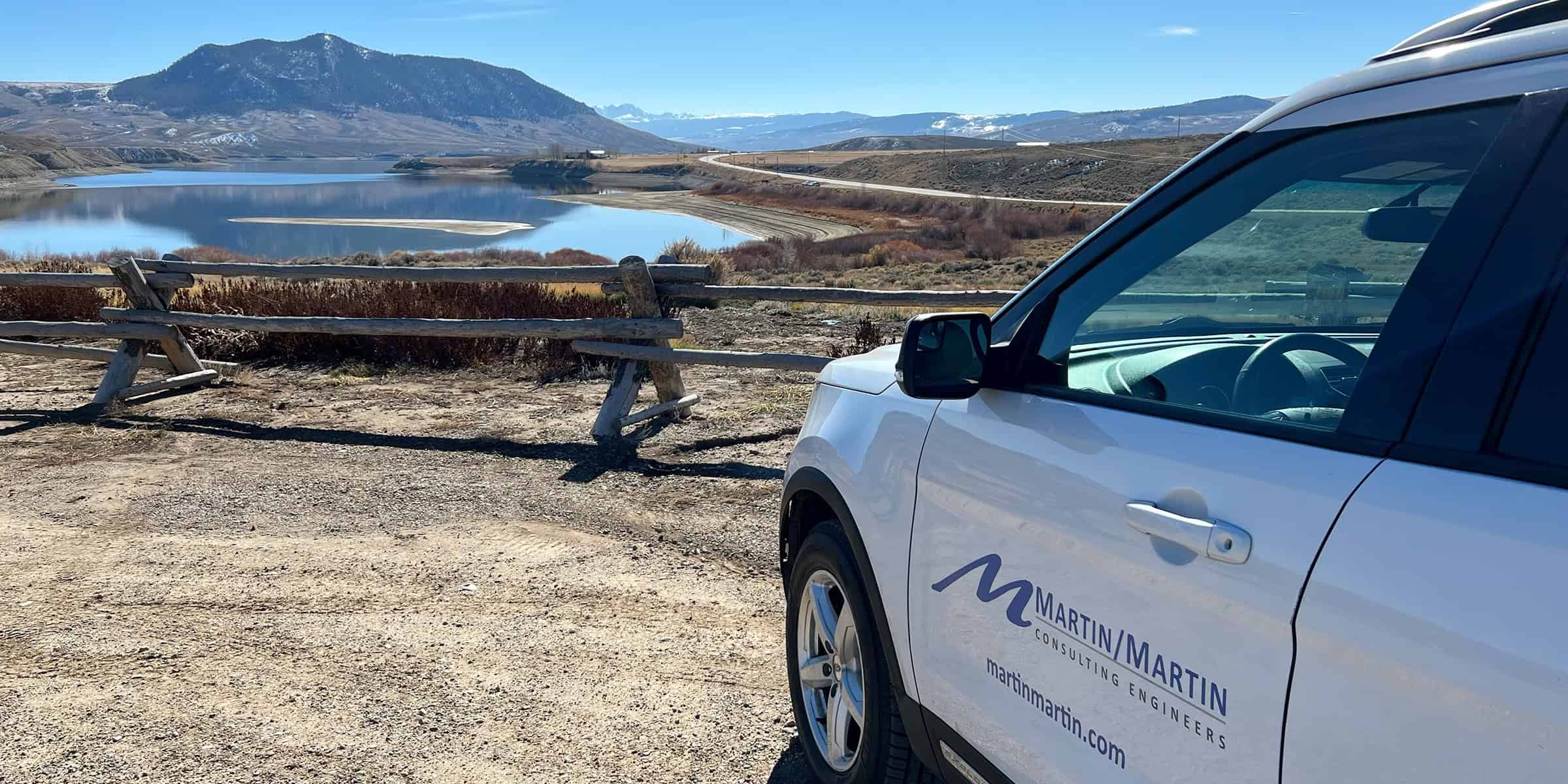 A white Martin/Martin company SUV is parked at an overlook, angled right, beside a split-rail wooden fence, overlooking a calm reservoir and distant snow-dusted mountains under a clear blue sky.

Text: MARTIN/MARTIN
CONSULTING ENGINEERS
martinmartin.com