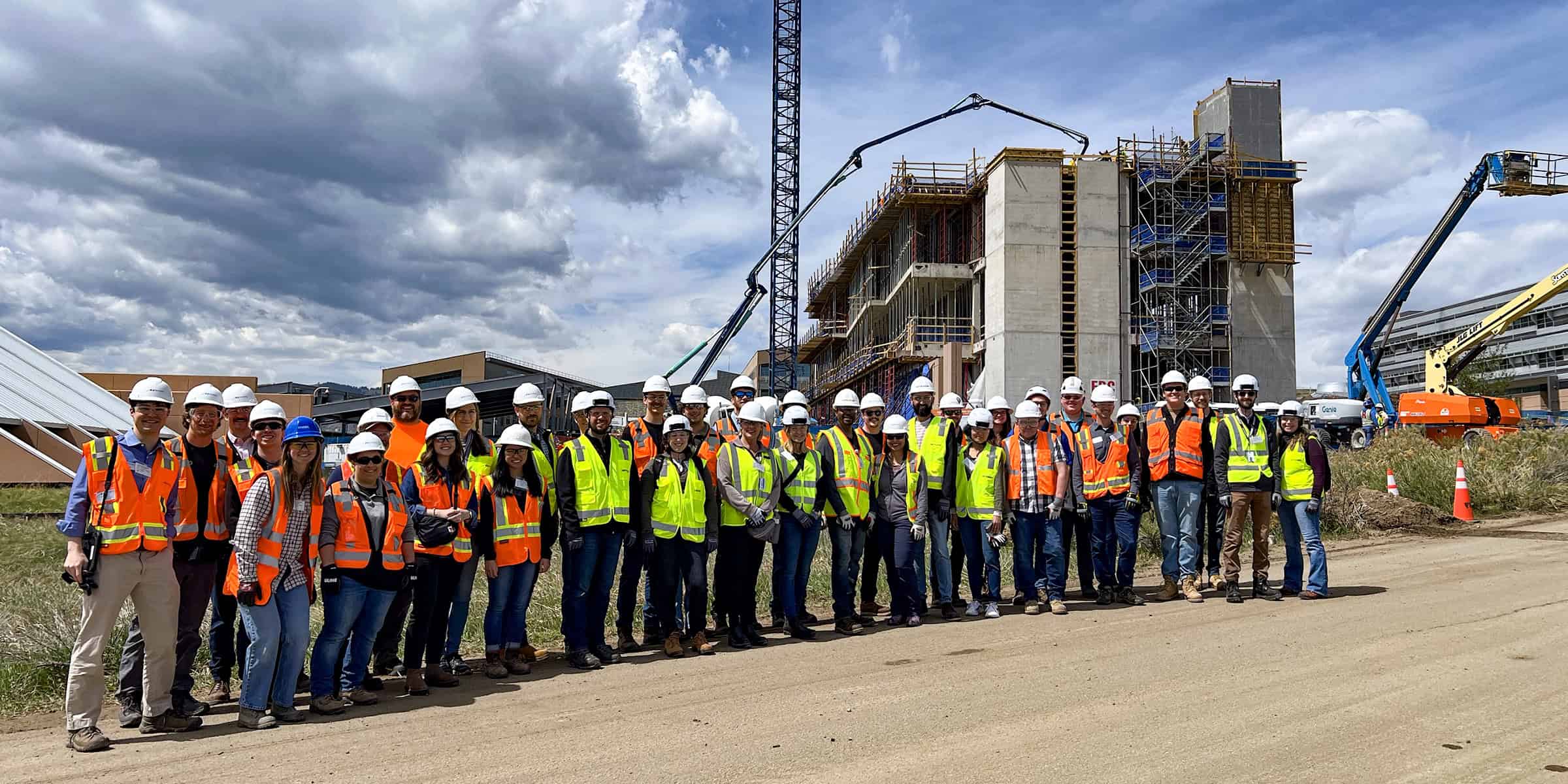 Large group of construction workers in hard hats and high-visibility vests pose on a dirt road before a multi-story concrete building under construction with cranes and cloudy sky. Text: "Genie"