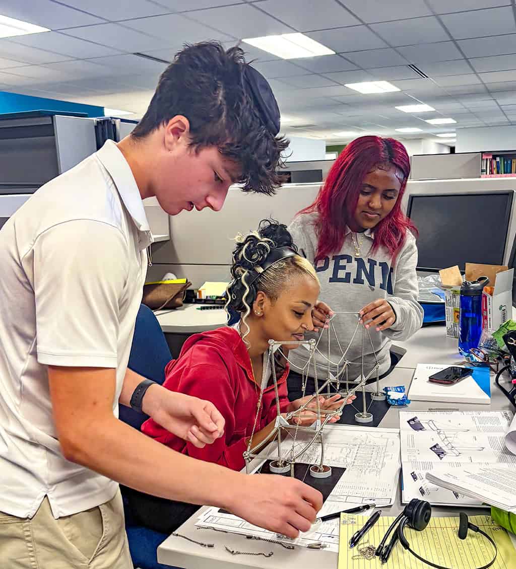 Small chain-and-post bridge model — three students assemble and adjust its cables — at a cluttered desk with papers, diagrams, headphones and cubicle workspace. Visible text: "PENN".