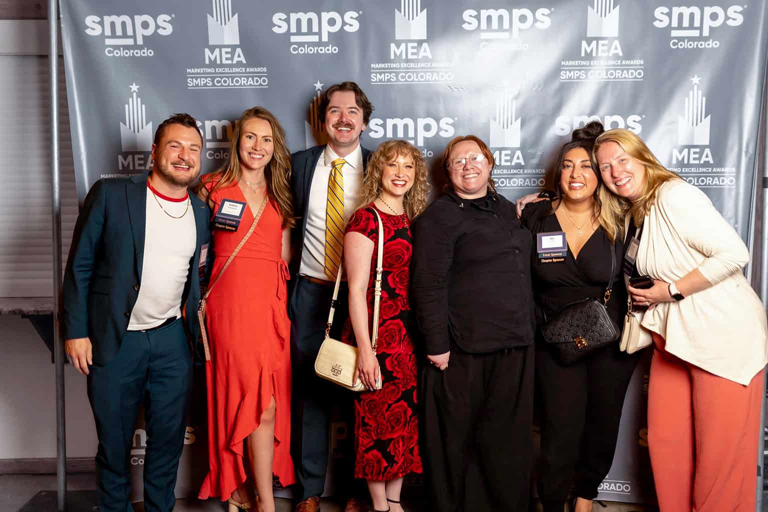 Group of seven adults standing close, smiling and posing for a photo in front of an event step-and-repeat backdrop.

Transcribed text:
- smps
- Colorado
- MEA
- MARKETING EXCELLENCE AWARDS
- SMPS COLORADO
- Event Sponsor
- Chapter Sponsor