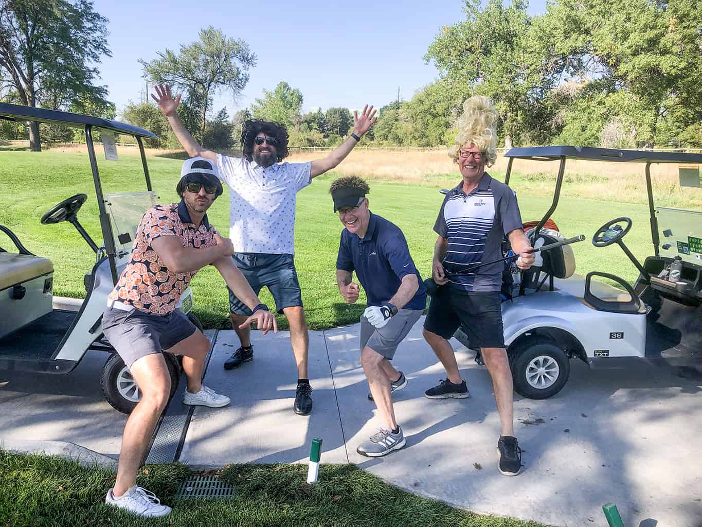 Four men striking playful poses—one wearing a tall blonde wig—gesturing and holding a golf club beside two golf carts on a sunlit fairway with trees and grassy rough.