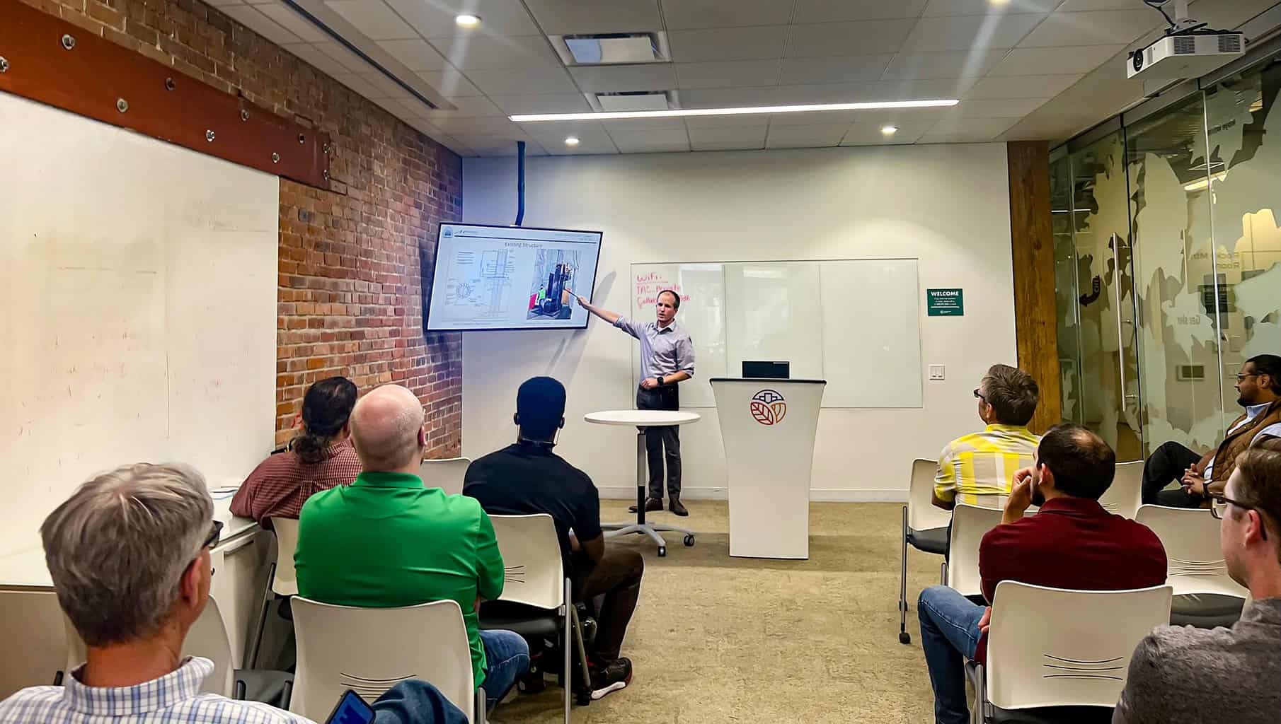 Presenter points at a wall-mounted screen while seated attendees watch in a small conference room with exposed brick, whiteboards, a podium, and a glass partition.

Text visible: "WiFi:" (pink marker on whiteboard); green sign: "WELCOME". Other marker writing is illegible.