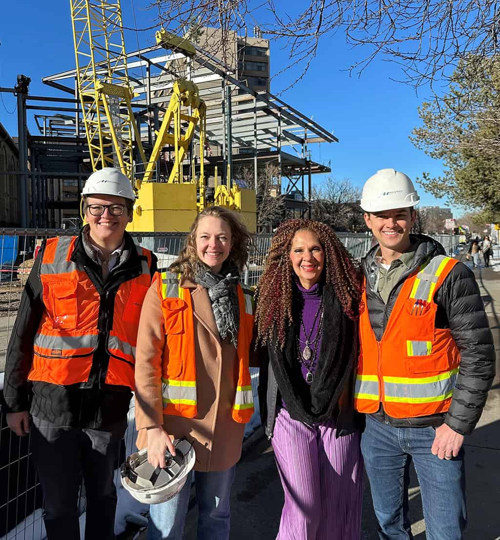 Four people wearing hard hats and orange safety vests stand smiling in front of an active construction site with a yellow crane and steel-frame structure under a clear blue sky.