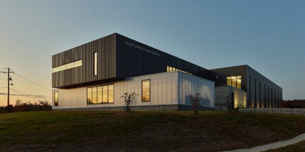 Modern school building labeled "VALLEY SPRINGS HIGH SCHOOL" (object) glows from interior lights (action) atop a grassy slope at dusk, framed by utility poles and a clear evening sky (context).