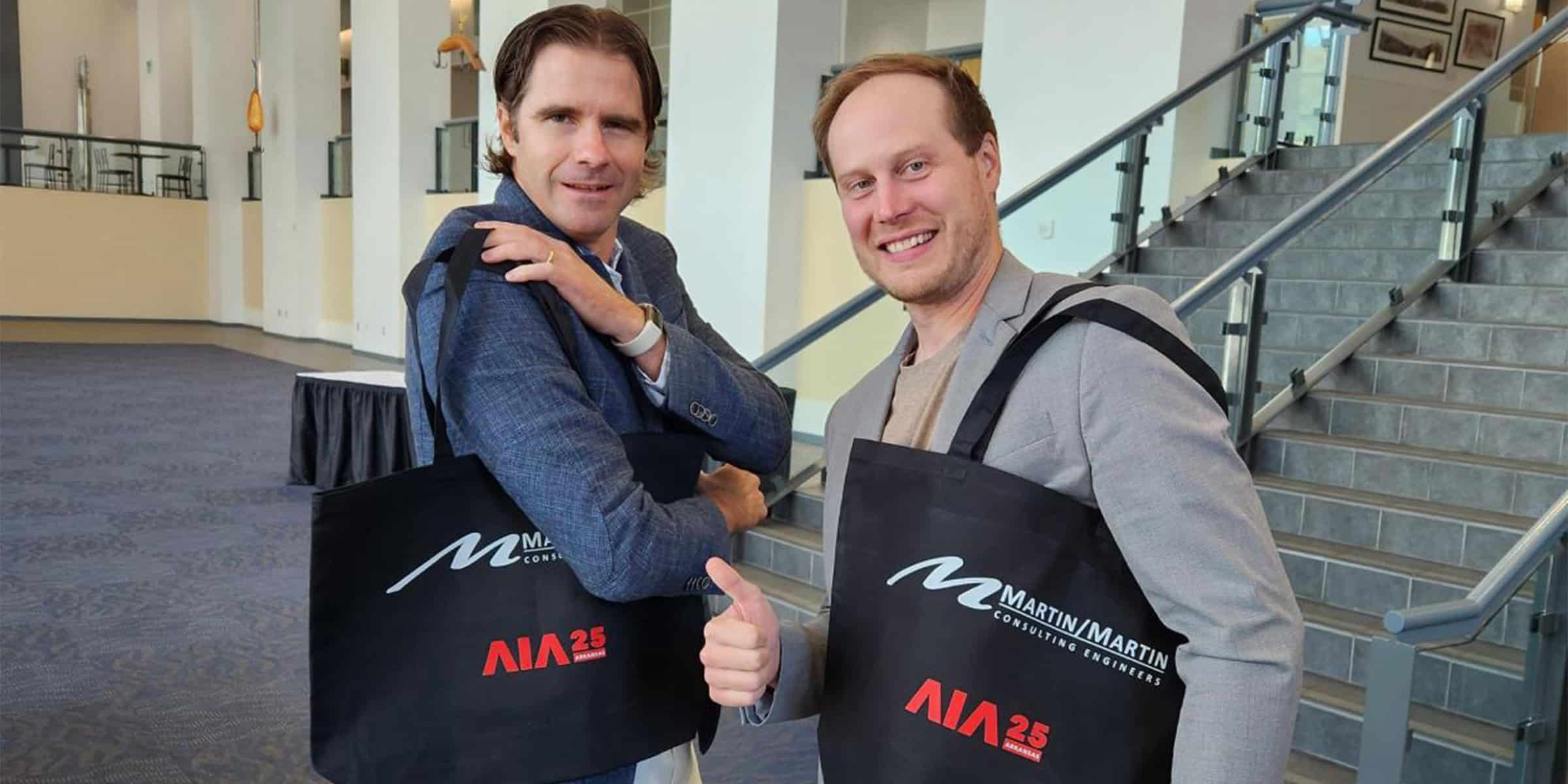 Two men stand smiling and posing with black conference tote bags; one gives a thumbs-up. They are in a carpeted convention lobby beside a tiled staircase.

Text in image: "MARTIN/MARTIN CONSULTING ENGINEERS" and "AIA 25 ARKANSAS"