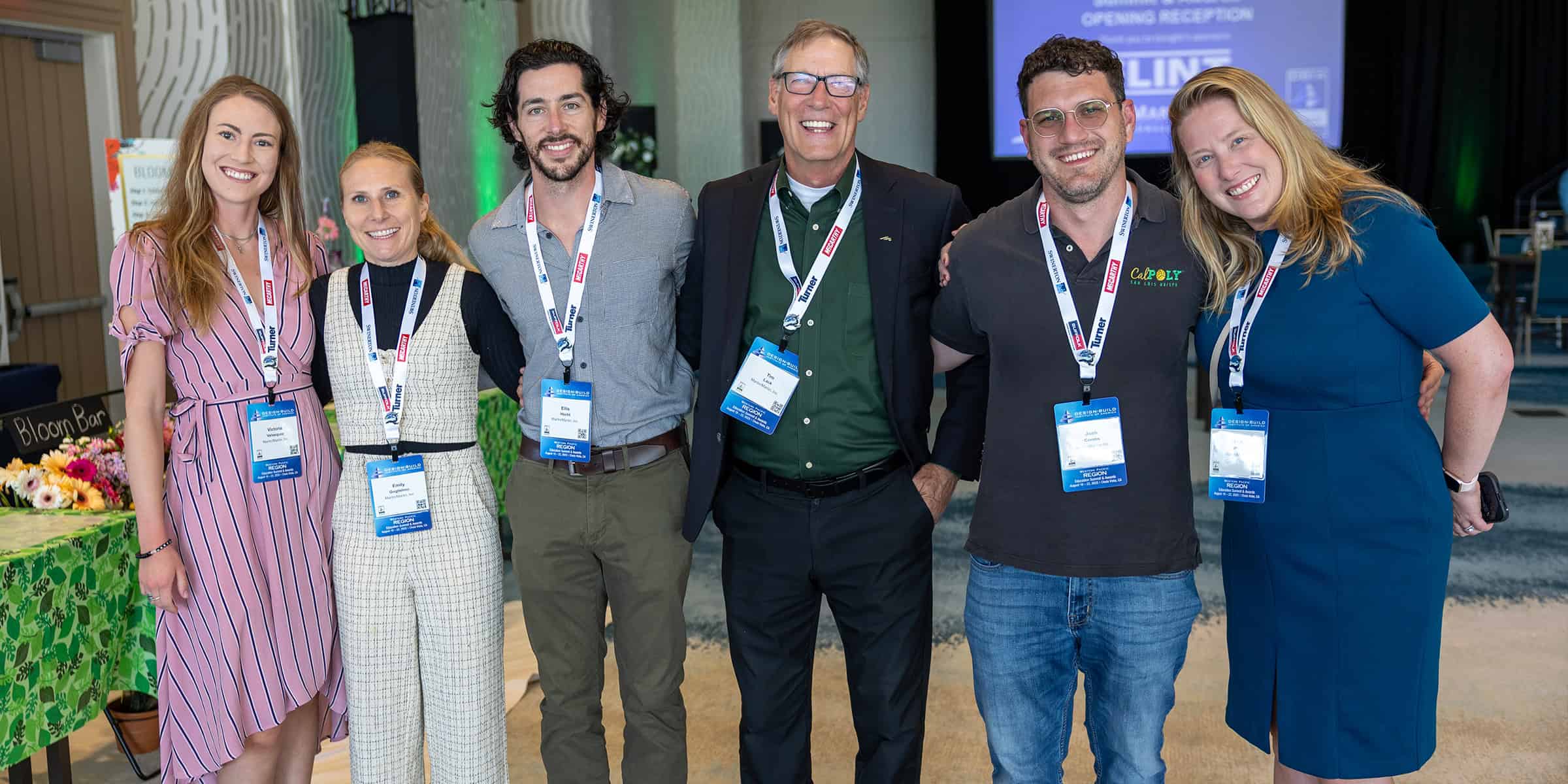 Group of six adults standing arm‑in‑arm, smiling at the camera, wearing conference lanyards in a carpeted event space with a flower table nearby.

Text visible: 
- Bloom Bar
- OPENING RECEPTION
- Cal Poly (on polo shirt)
- Turner (on lanyards)
- EXHIBITOR (on red lanyard tabs)
- REGION (on badge ribbons)