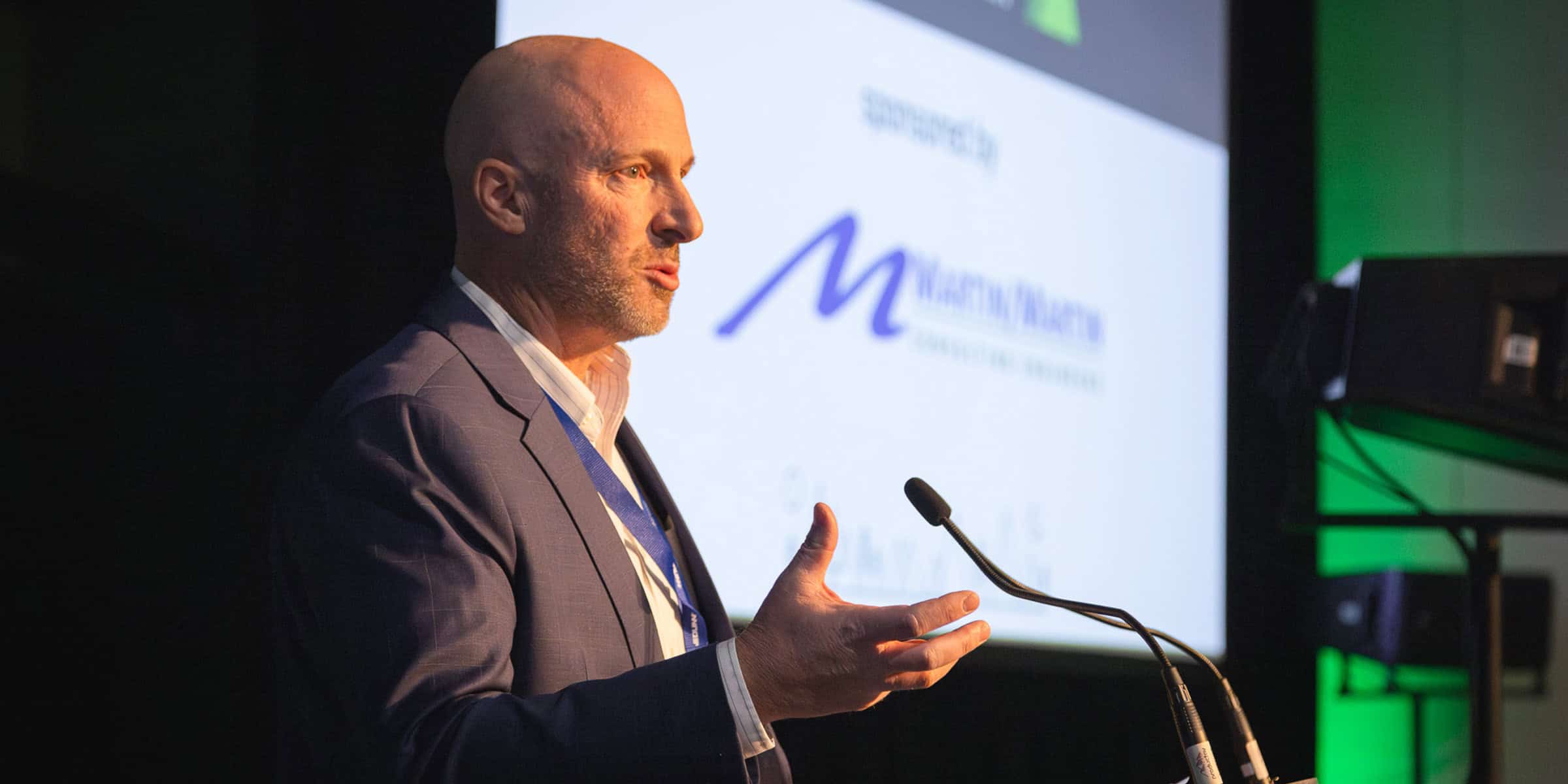 Bald man gestures while speaking at a podium before a projected slide in a dim conference room.

Text on screen: "sponsored by" ; stylized "M" logo ; "[company name illegible]" ; "CONSULTING SERVICES."