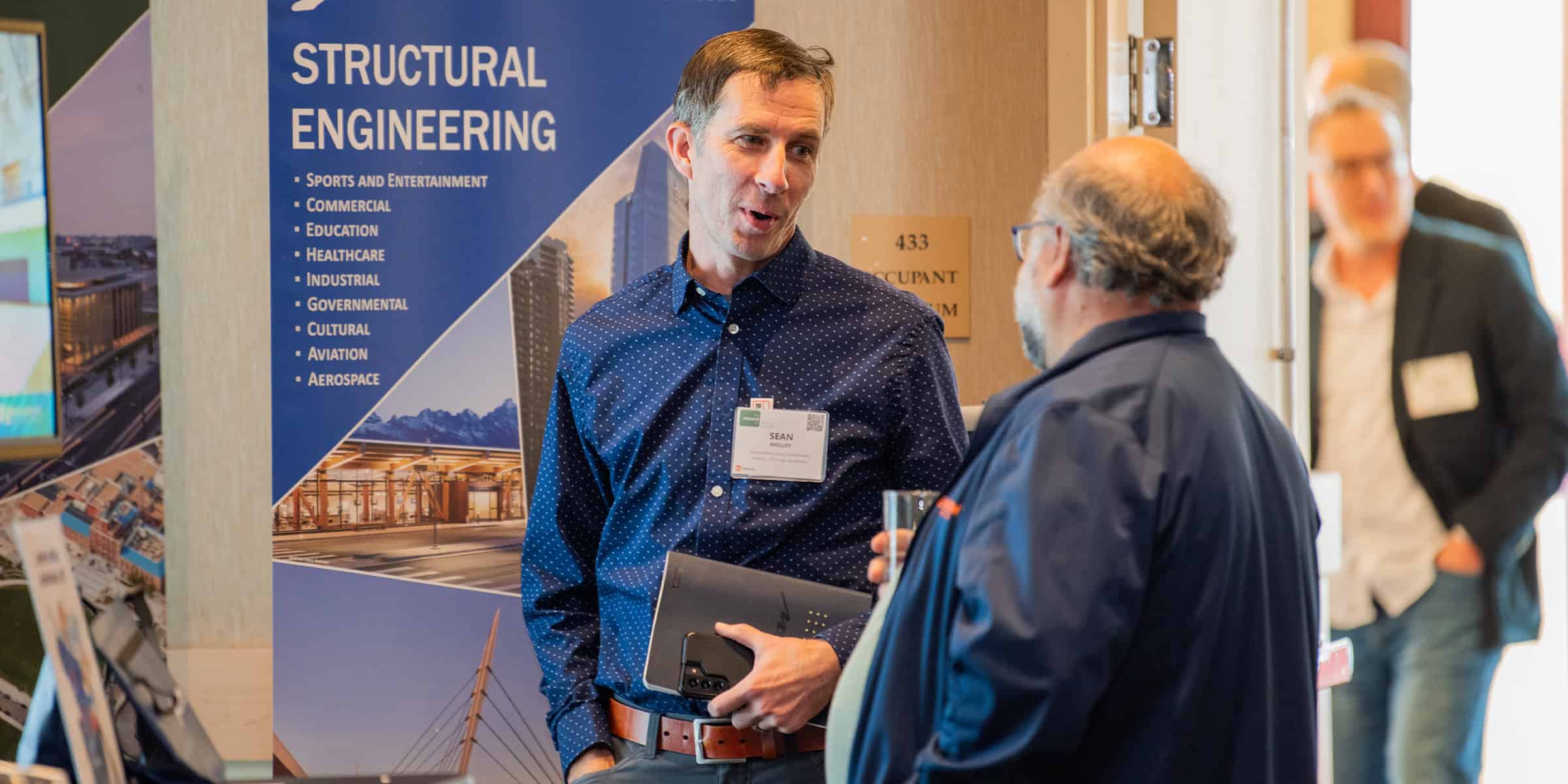 A man in a blue patterned shirt holds a tablet and phone while talking to another attendee at a conference booth, with a structural engineering banner and room doorway behind them.

Transcribed text:
- STRUCTURAL ENGINEERING
- Sports and Entertainment
- Commercial
- Education
- Healthcare
- Industrial
- Governmental
- Cultural
- Aviation
- Aerospace
- 433
- OCCUPANT
- SEAN