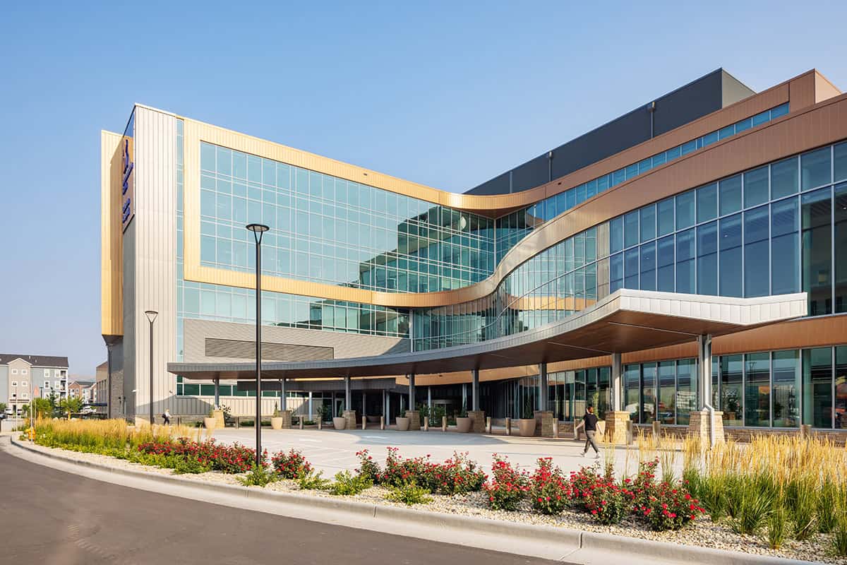 Modern glass-and-metal building forms a sweeping curved façade and covered entrance; a few pedestrians walk by as landscaped flowerbeds and ornamental grasses line the driveway under a clear blue sky.