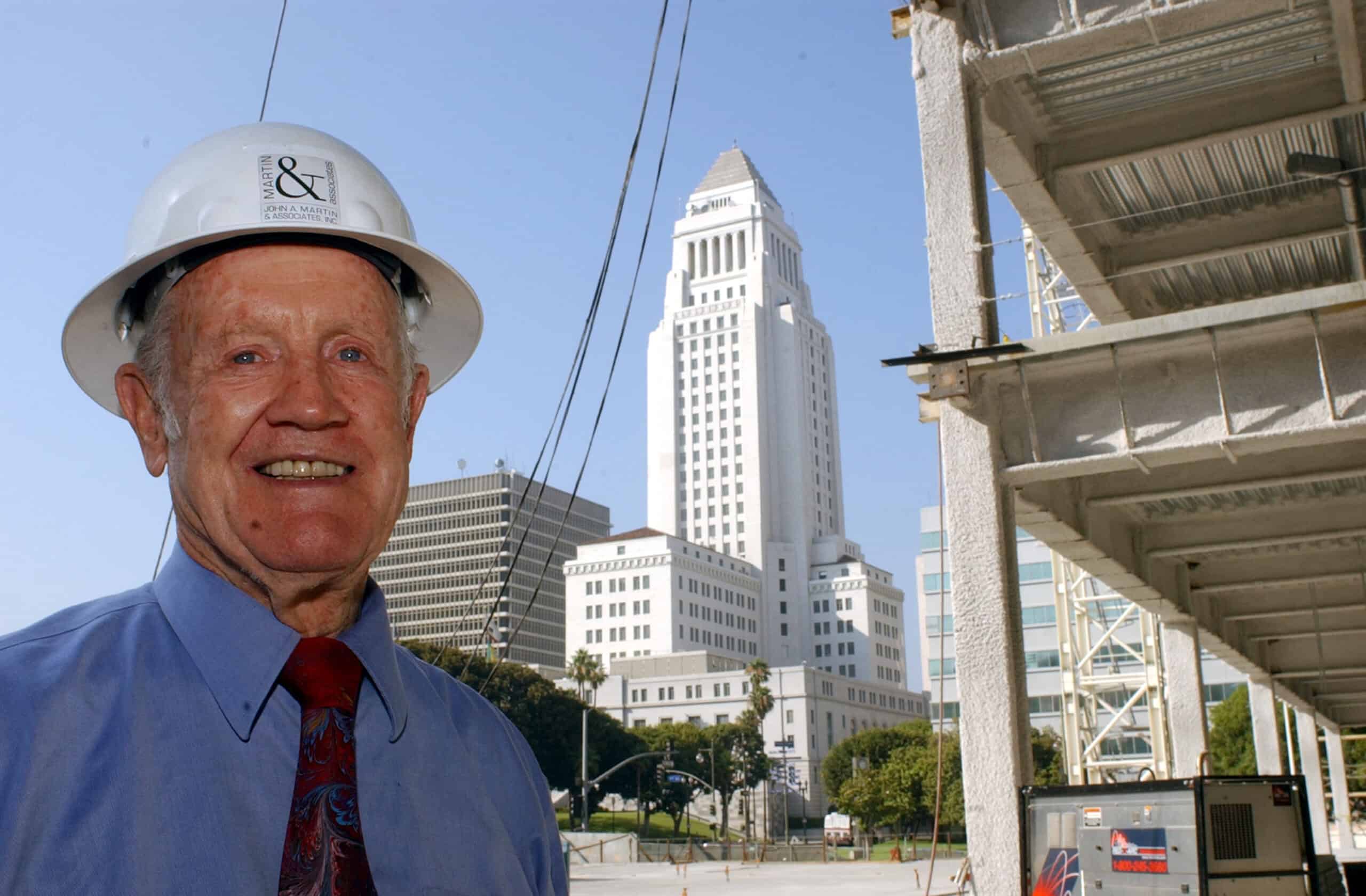 An older man wearing a white hard hat smiles at the camera, standing at a construction site beside exposed concrete framing, with a tall classical city tower and office buildings behind him.

Text on hard hat: "MARTIN &" and "JOHN A. MARTIN & ASSOCIATES, INC." (other small equipment text not legible)