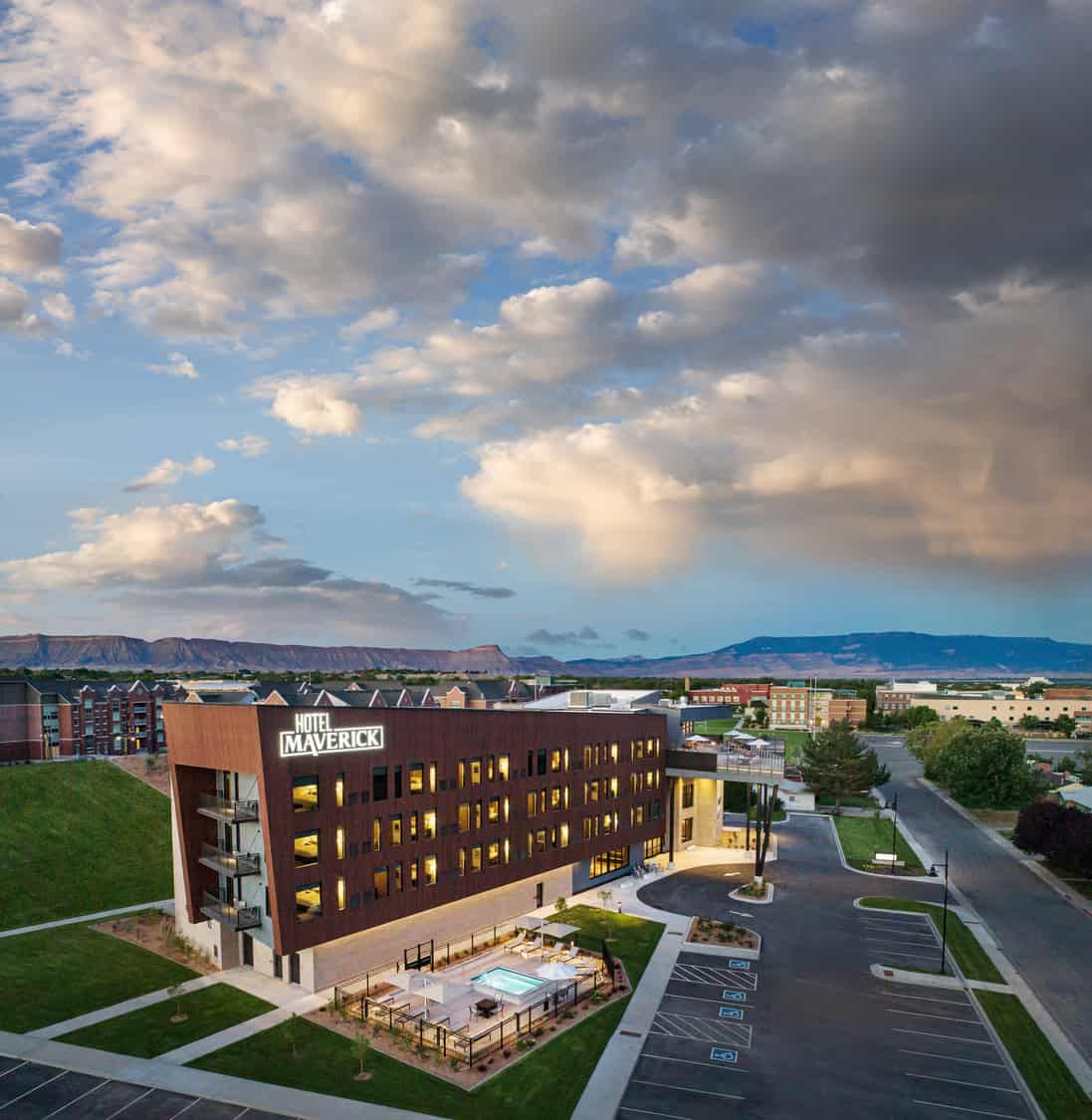 Hotel Maverick building stands illuminated at dusk, windows and outdoor pool glowing; surrounded by empty parking, lawns and distant mesas beneath a dramatic, cloud-filled sky. Text: "HOTEL MAVERICK".