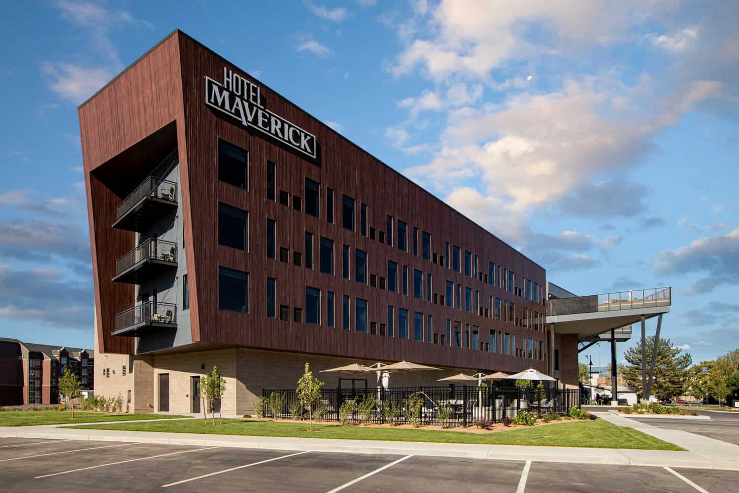 Hotel Maverick (sign: "HOTEL MAVERICK"): angular wood-clad building displaying recessed balconies and ground-level patio with umbrellas, sitting beside a parking lot and lawn under a blue, partly cloudy sky.