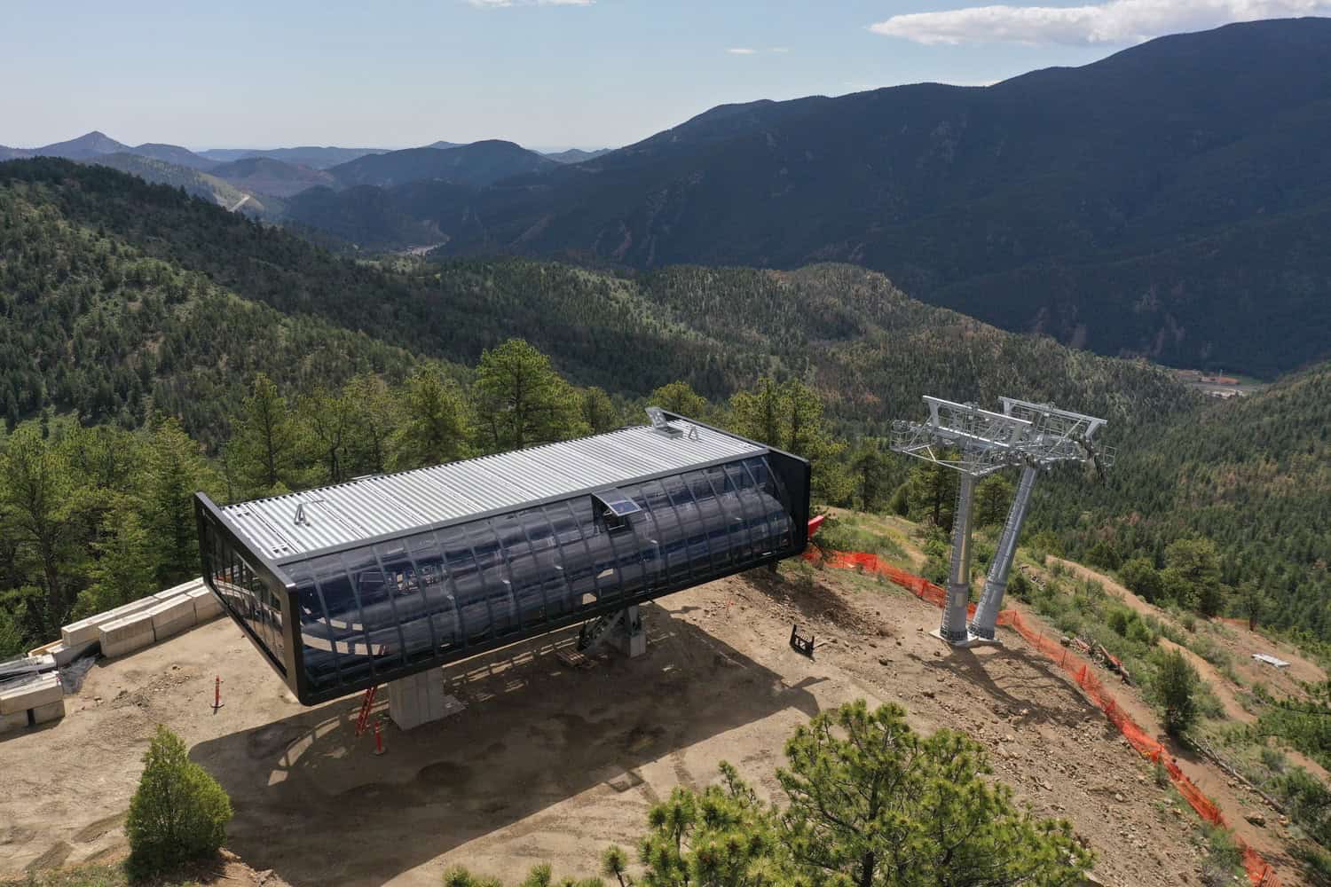 Ski-lift terminal sits on a cleared mountain slope, connected to a steel support tower, overlooking dense pine forests and layered ridgelines of a deep, distant mountain valley.