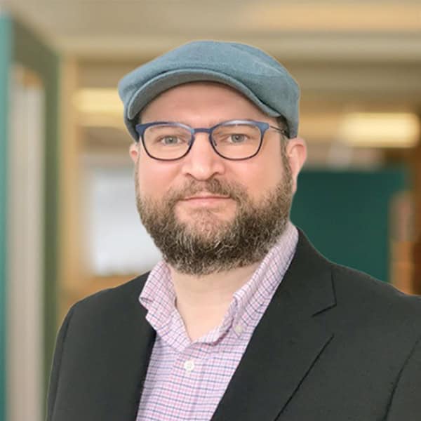Bearded man wearing a blue flat cap and glasses faces the camera with a mild smile, dressed in a black blazer and checkered shirt, standing in a blurred office corridor.