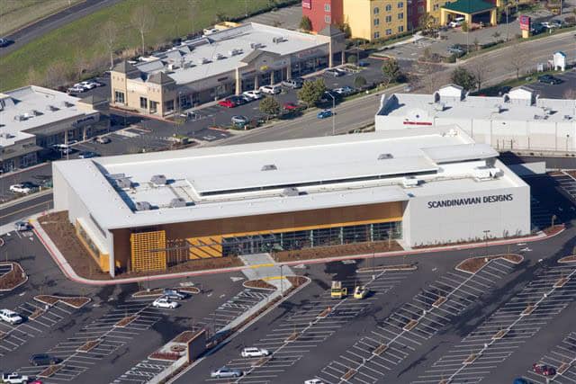 A large rectangular retail building labeled "SCANDINAVIAN DESIGNS" sits amid a mostly empty parking lot in a suburban shopping center, shown from an aerial perspective.