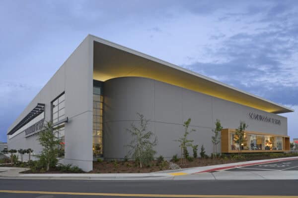 Large modern retail building displays interior lighting and showrooms through glass, sitting amid landscaped beds and a roadway at dusk. Text: SCANDINAVIAN DESIGNS