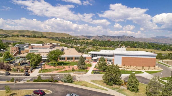 Modern brick-and-glass campus building sits amid lawns and walkways, playground to the left, parked cars in the foreground, and rolling foothills beneath a blue sky with scattered clouds.