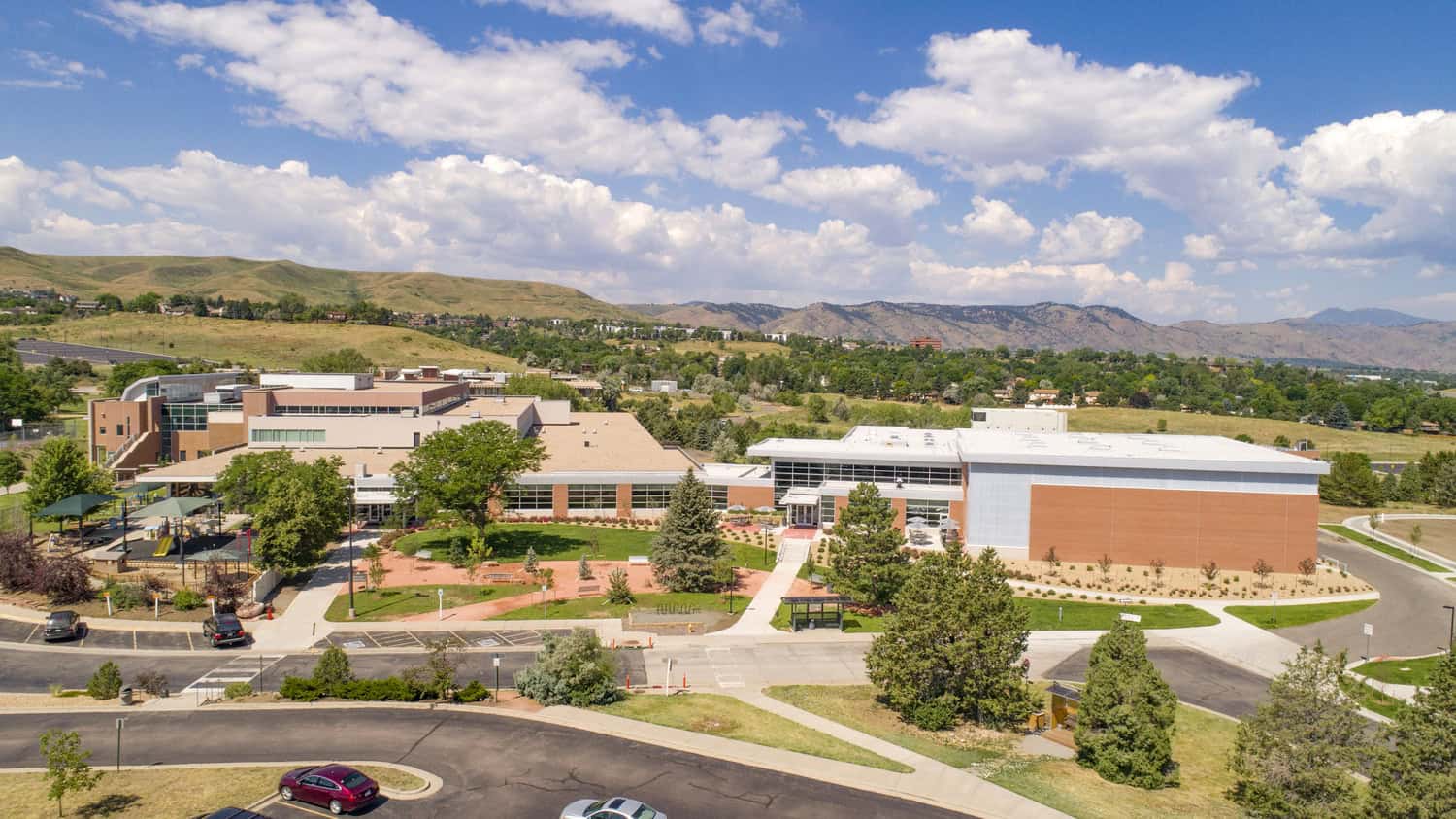 Modern brick-and-glass campus building sits amid lawns and walkways, playground to the left, parked cars in the foreground, and rolling foothills beneath a blue sky with scattered clouds.
