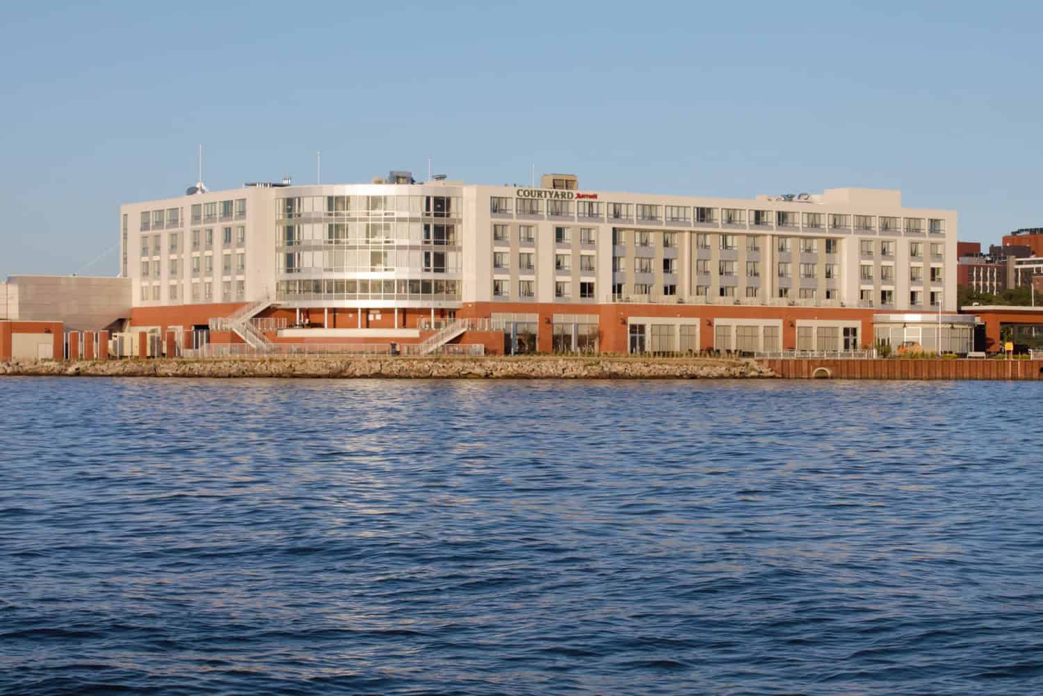 A five-story waterfront hotel sits on a rocky quay, sunlight glinting on its glass facade; calm blue harbor waters ripple in front, clear sky overhead. Text on building: "COURTYARD Marriott".