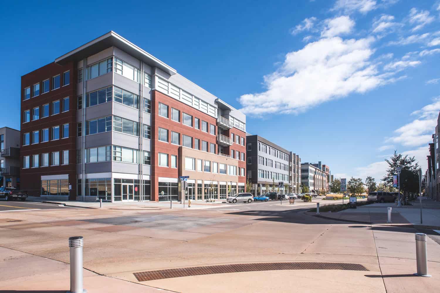 A five-story mixed-use brick-and-glass building faces a sunny urban intersection, with parked cars, bollards, and a clear blue sky; visible sign reads "20".