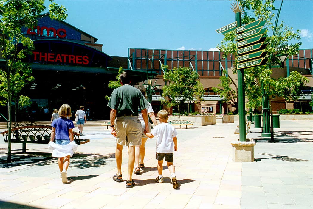 Family of four (two adults, two children) walk toward a red‑lettered cinema entrance across a sunny, tree‑lined plaza with benches and a tall directional sign.

Text found in the image:
- amc (logo on building)
- THEATRES (marquee)
- Directional sign (top to bottom): AMC THEATRES; SHOPS; RESTAURANTS; BRIDGE; ICE CENTRE; OFFICES; WESTIN HOTEL
- Other small posters near the entrance are present but not legible.